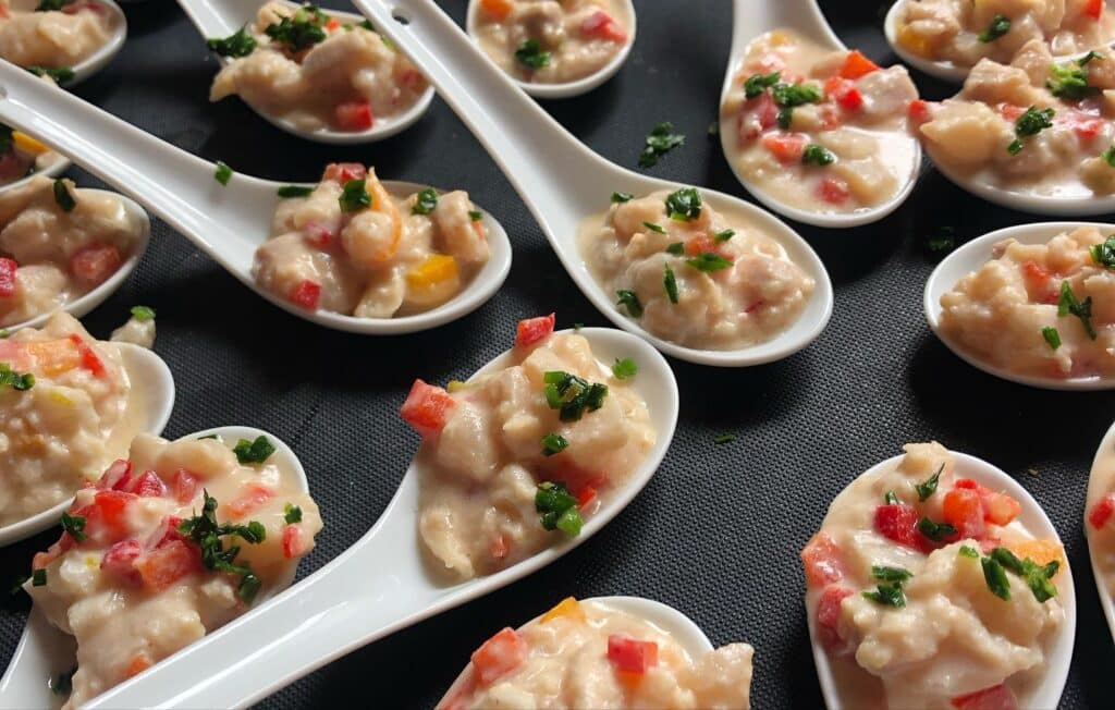 Amuse-bouche fruits de mer : verrines cocktail festif Amuse-bouche de fruits de mer crémeux et dés de poivrons rouges et verts, servis en cuillères de dégustation blanches sur fond noir.