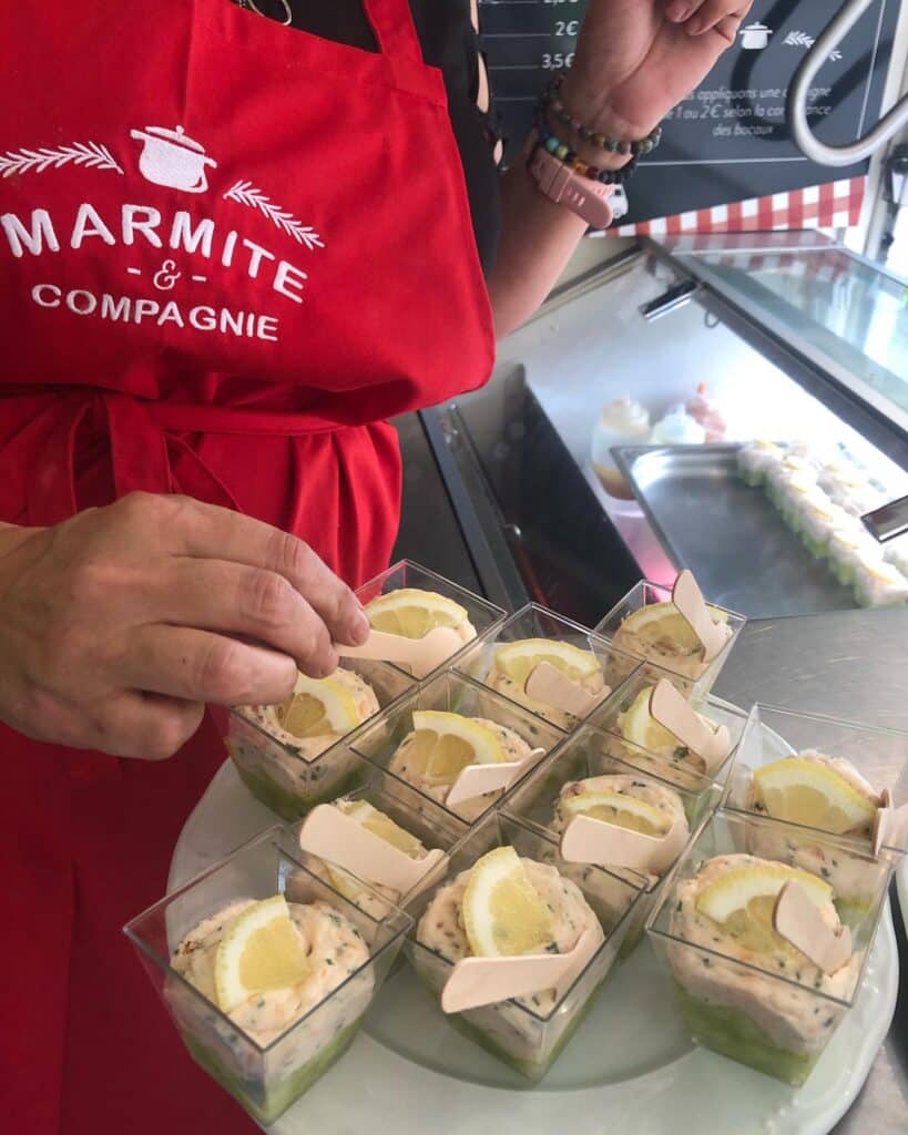Verrines Apéritives Marmite & Compagnie : Présentation Fraîche Employé Marmite & Compagnie en tablier rouge dressant des verrines apéritives saumon-citron, avec un menu affichant des prix.